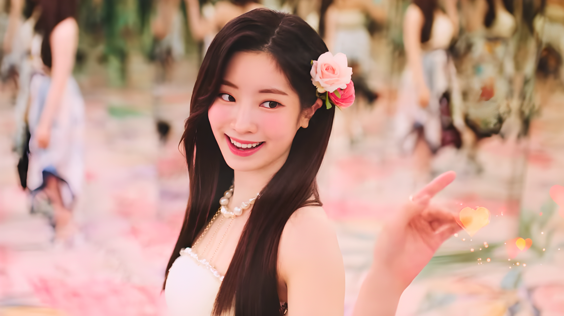 HD PC wallpaper of a smiling K-pop idol singer wearing a pink flower in her hair, radiating summer vibes amid a soft pastel, floral-filled background.