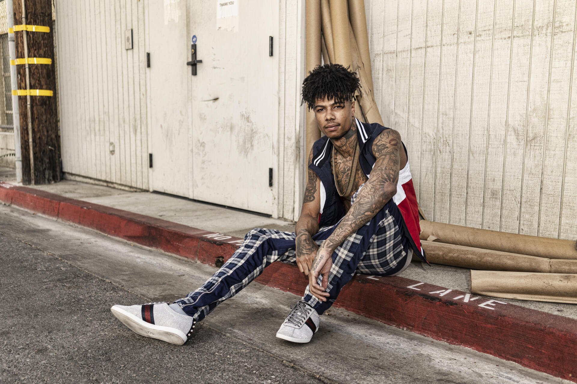 Tattooed rapper in plaid pants and sneakers sits on a curb by a warehouse door, framed as a 5K Ultra HD PC desktop wallpaper/background.