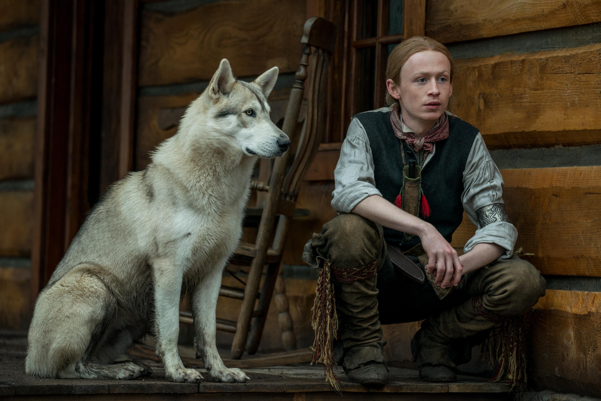Outlander TV scene: a young man seated beside a Siberian husky in a rustic cabin, 5K Ultra HD PC desktop wallpaper.