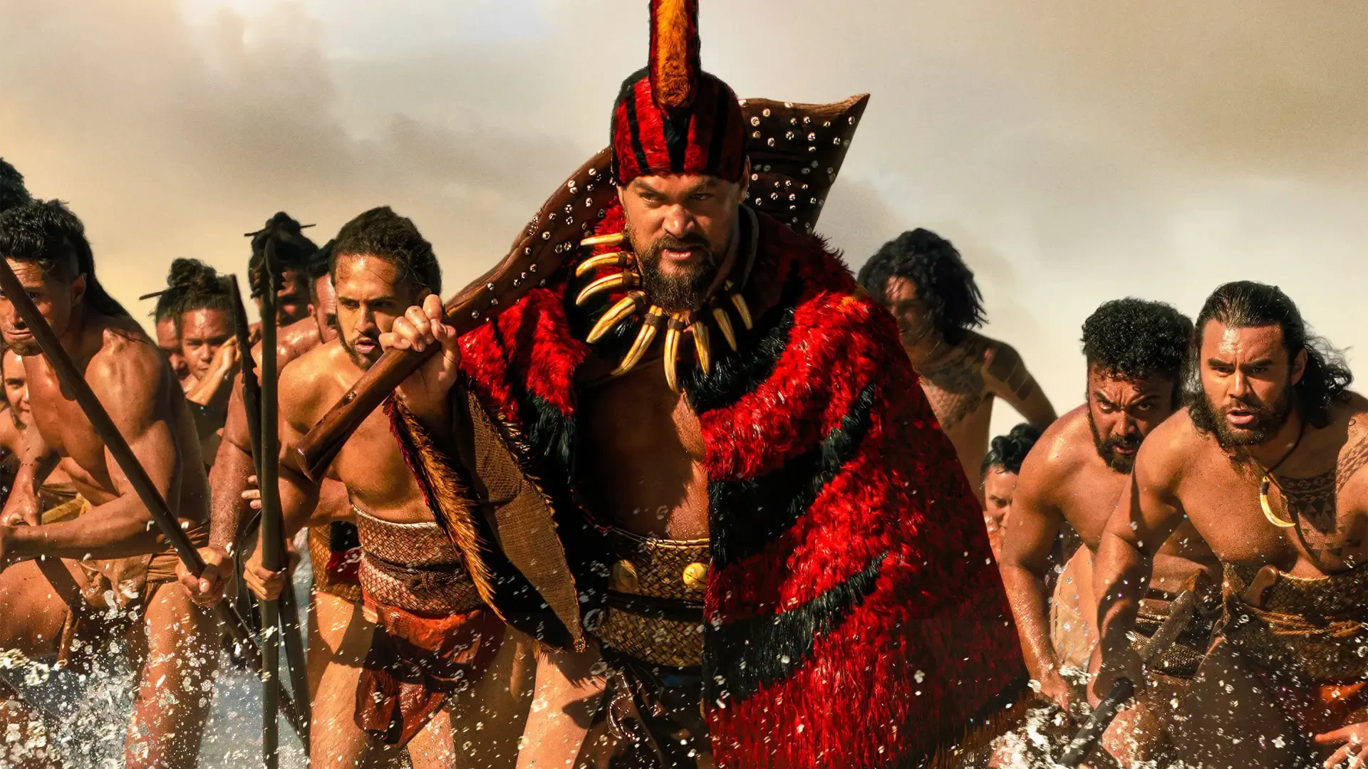 Chief of War leads a charging warband through surf, clad in a red feathered cloak and tooth necklace, warriors with spears behind — 4K Ultra HD PC desktop wallpaper/background.