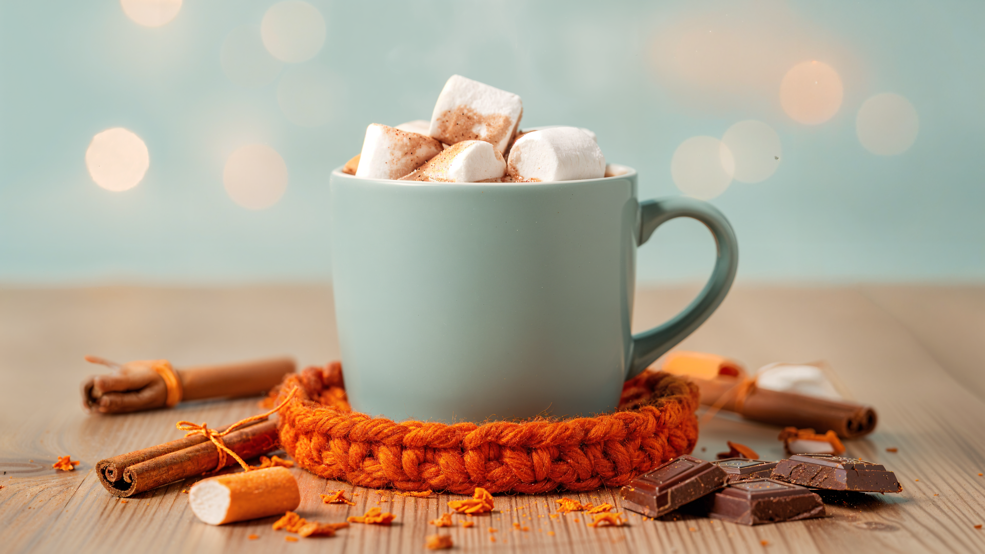 4K Ultra HD PC desktop wallpaper: steaming mug of hot cocoa with marshmallows on an orange knit coaster, cinnamon sticks and chocolate pieces on a wooden table, soft bokeh background.