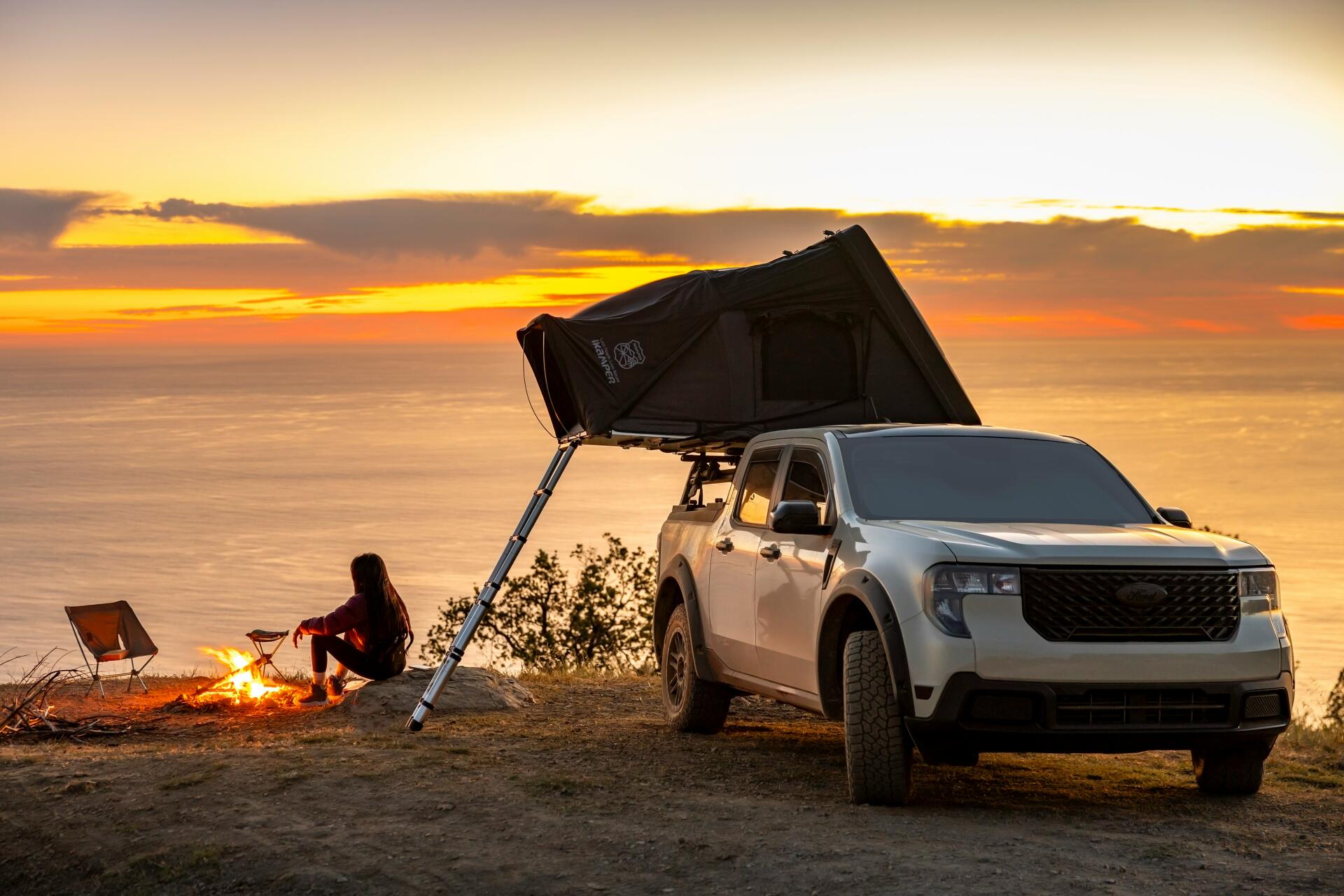 5K Ultra HD PC desktop wallpaper: Ford Maverick pickup at a camping spot with rooftop tent and morning bonfire on a coastal bluff, camper silhouetted against sunrise.