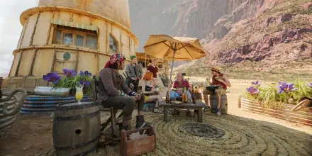 2K Quad HD PC desktop wallpaper of a TV show scene from One Piece (Live Action): Straw Hat crew relaxing around tables and umbrellas outside a desert tavern by rocky cliffs.