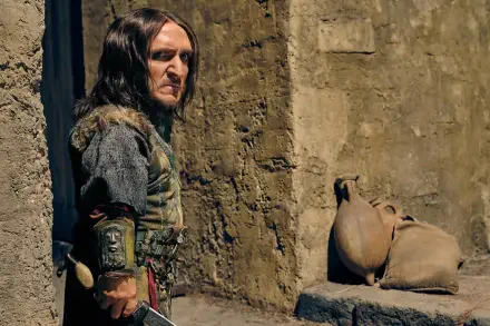 HD PC desktop wallpaper: a dwarf character from the TV show Spartacus: House of Ashur in leather armor stands in a sunlit stone courtyard beside clay jars.