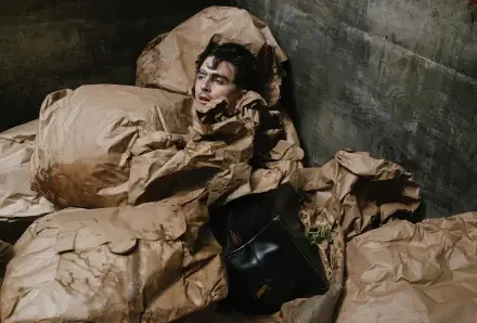 2K Quad HD movie still (Marty Supreme) desktop wallpaper: young man emerging from crumpled paper sacks in a dim concrete room, black bag beside him, weary expression.