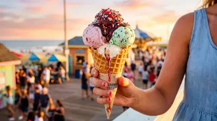 Close-up of a hand holding a triple-scoop, sprinkle-topped waffle ice cream cone at a bustling boardwalk sunset — vivid 5K Ultra HD PC Desktop Wallpaper and Background.
