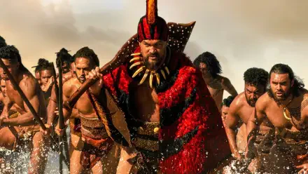 Chief of War leads a charging warband through surf, clad in a red feathered cloak and tooth necklace, warriors with spears behind — 4K Ultra HD PC desktop wallpaper/background.