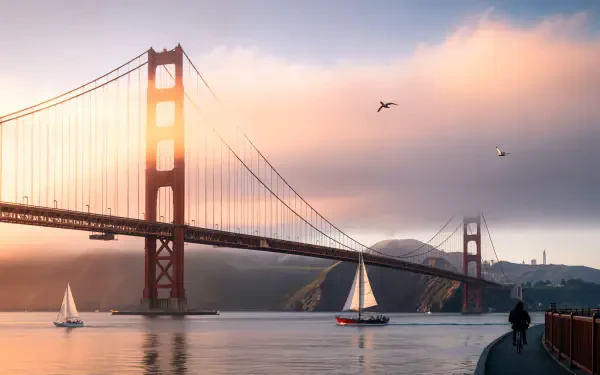 4K Ultra HD PC desktop wallpaper of the Golden Gate Bridge at sunrise, sailboats on calm water, soft pastel sky and a lone walker on the waterfront.