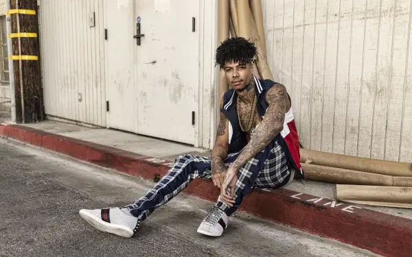 Tattooed rapper in plaid pants and sneakers sits on a curb by a warehouse door, framed as a 5K Ultra HD PC desktop wallpaper/background.