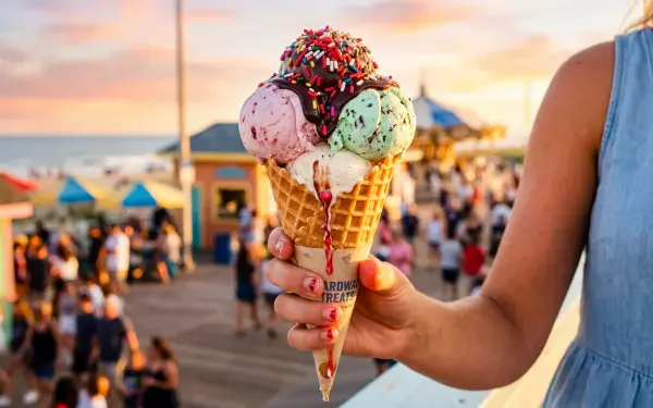 Close-up of a hand holding a triple-scoop, sprinkle-topped waffle ice cream cone at a bustling boardwalk sunset — vivid 5K Ultra HD PC Desktop Wallpaper and Background.