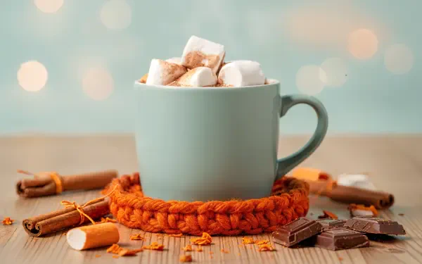 4K Ultra HD PC desktop wallpaper: steaming mug of hot cocoa with marshmallows on an orange knit coaster, cinnamon sticks and chocolate pieces on a wooden table, soft bokeh background.
