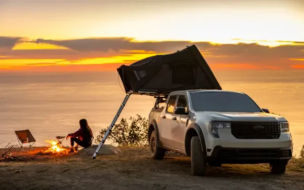 5K Ultra HD PC desktop wallpaper: Ford Maverick pickup at a camping spot with rooftop tent and morning bonfire on a coastal bluff, camper silhouetted against sunrise.