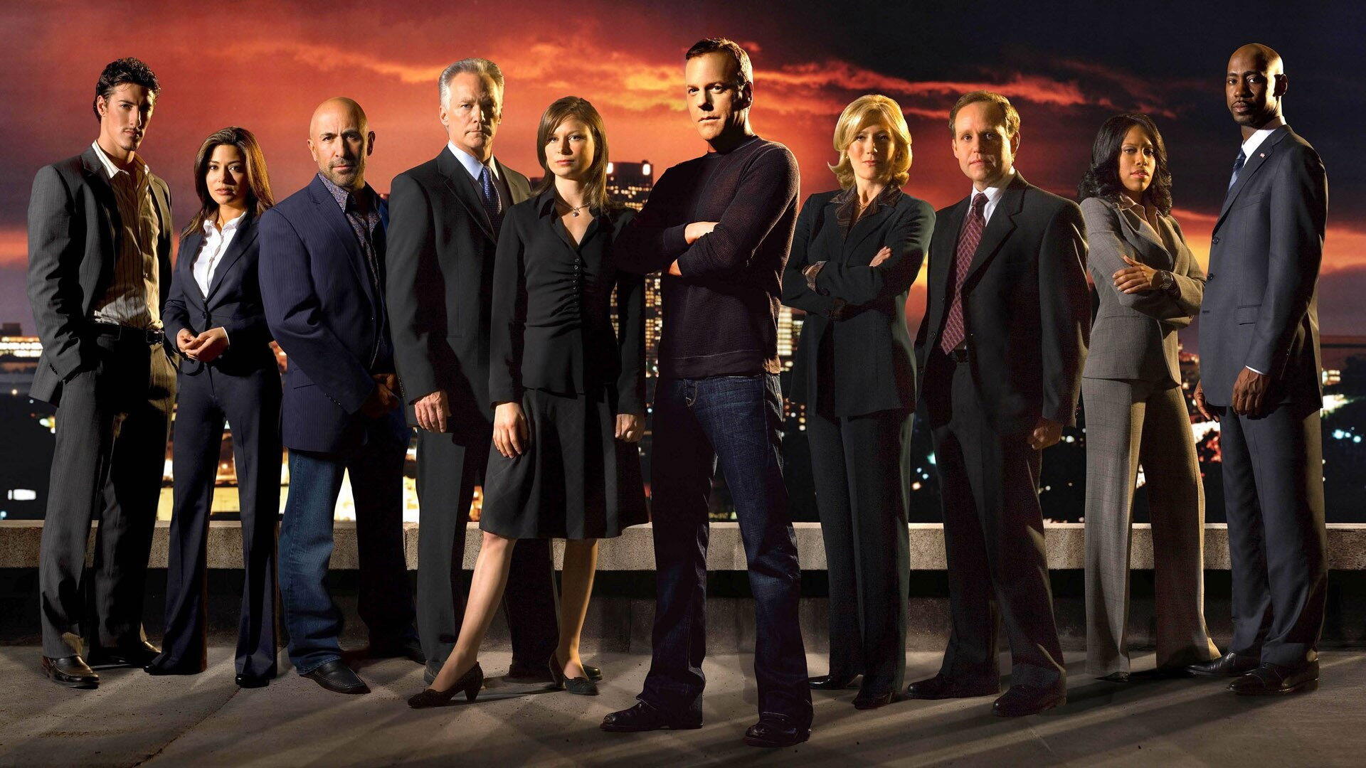 HD PC desktop wallpaper — 24: a group of serious men and women in professional attire stand on a rooftop before a fiery sunset skyline, a central figure posed with arms crossed.