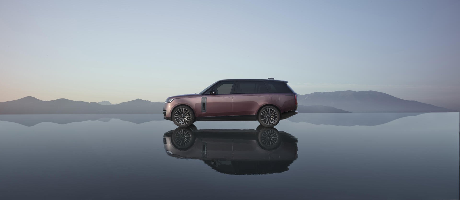 Range Rover Electric SUV in profile reflected on a mirror-like surface with distant mountains — 8K Ultra HD PC desktop wallpaper/background.