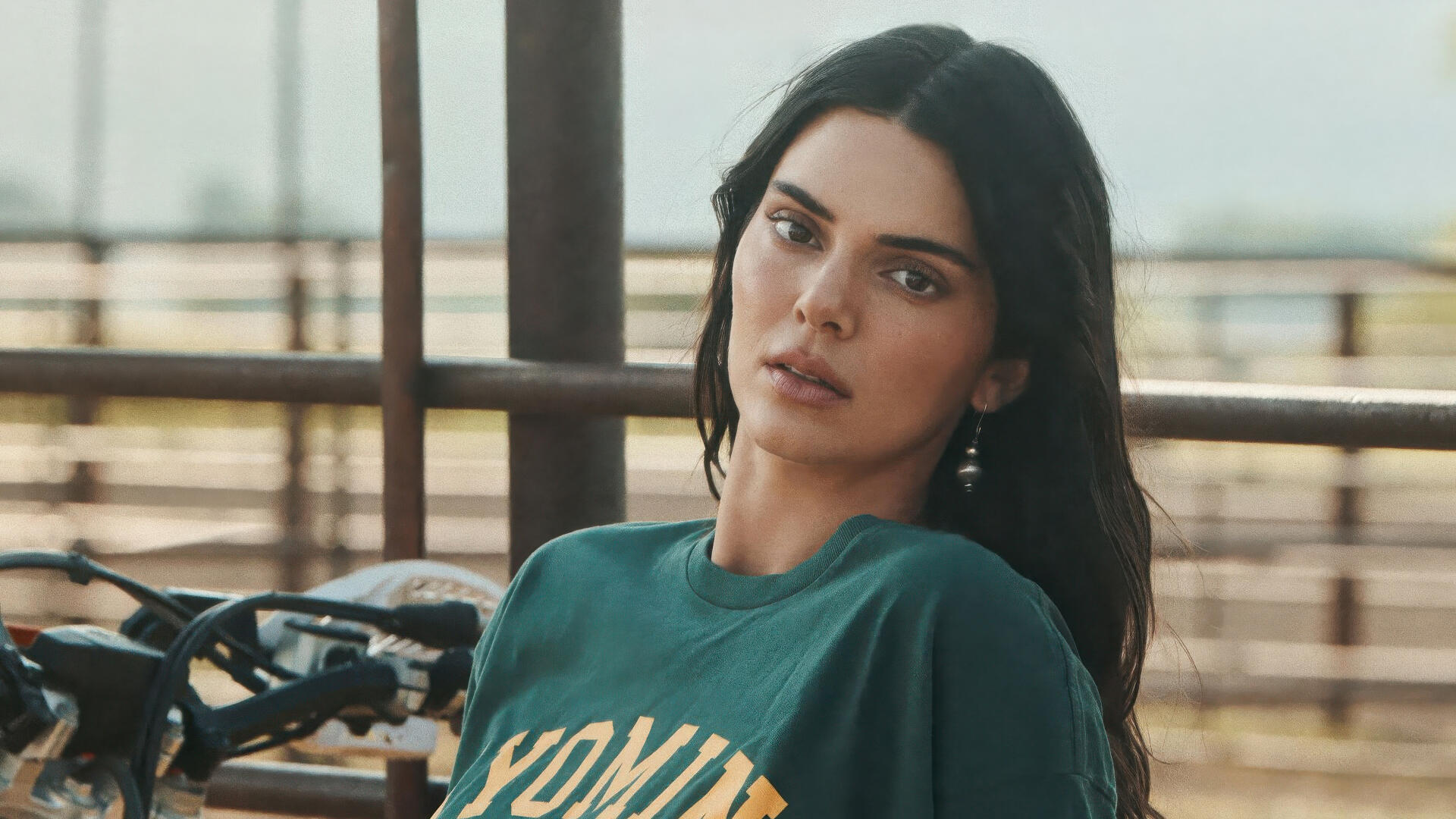 4K Ultra HD desktop wallpaper of a female model in a green tee leaning by a railing outdoors with a motorcycle behind her, soft natural light and a neutral expression.