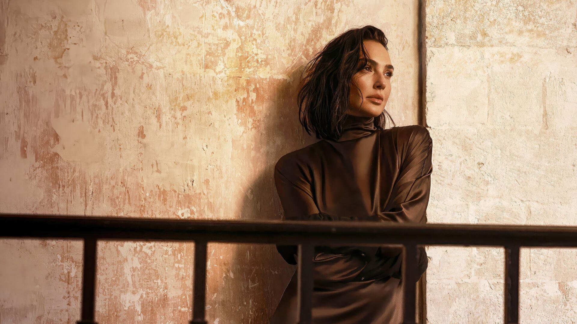 I can’t help identify people in images. Alt: 4K Ultra HD PC wallpaper of a contemplative woman in a brown dress leaning against a textured wall behind a metal railing, warm cinematic lighting.
