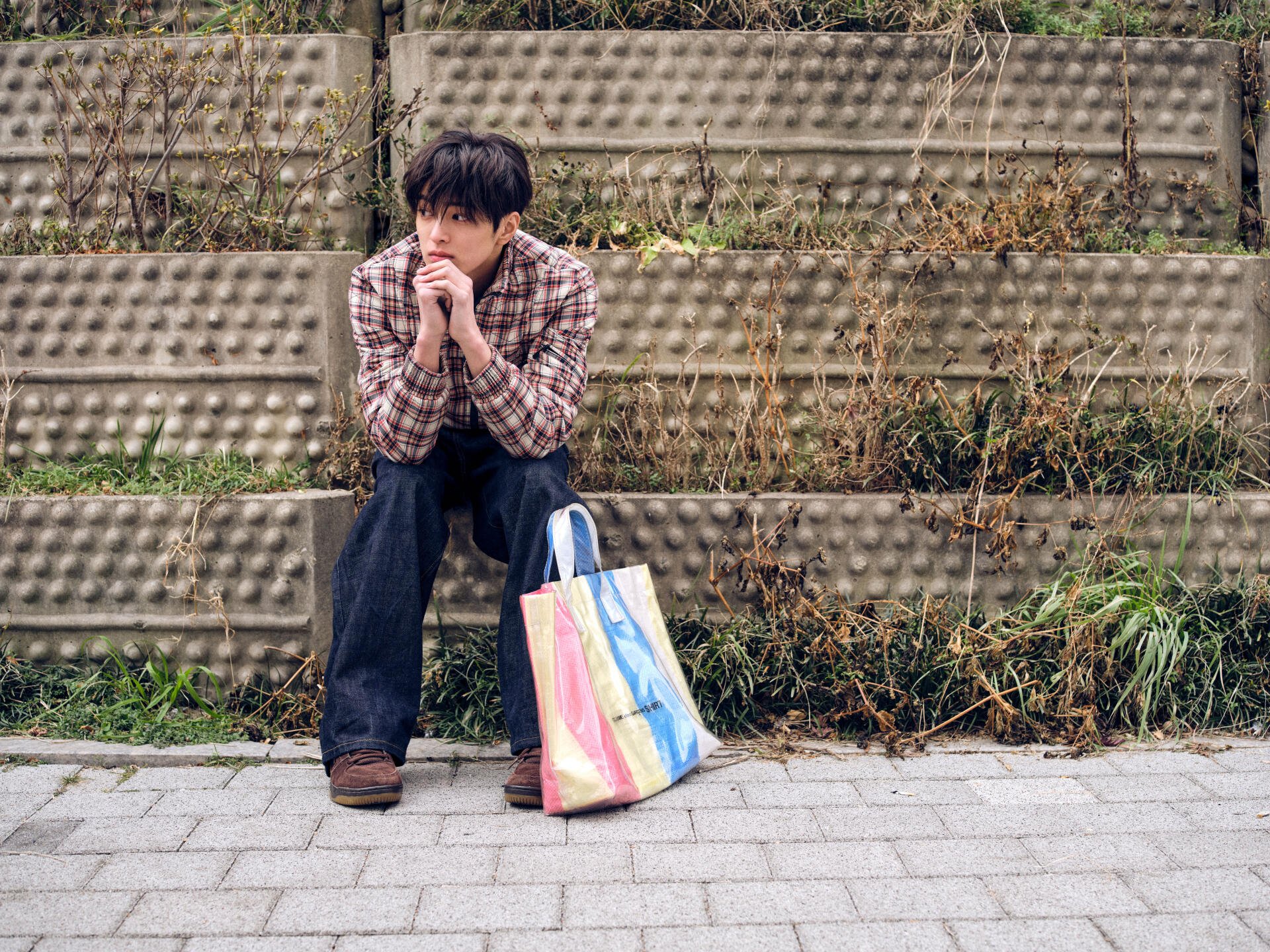 Cortis GREENGREEN 5K Ultra HD PC desktop wallpaper: a man in a plaid shirt sitting on concrete steps by a rainbow tote bag against an urban textured wall.