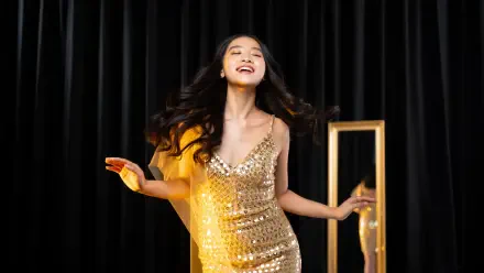 Cheerful young woman in a sparkling gold dress dancing before black curtains, light streaks and a reflected frame — 5K Ultra HD PC Desktop Wallpaper and Background.