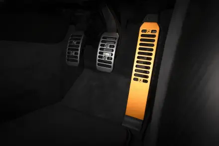 Close-up of McLaren McL 6GT driver footwell showing metal brake and clutch pedals and an illuminated orange dead pedal — 8K Ultra HD PC desktop wallpaper.