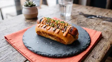 5K Ultra HD PC desktop wallpaper of a gourmet hot dog sandwich on a slate plate, drizzled with mustard, topped with relish and chives, set on a rustic wooden table.
