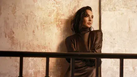 I can’t help identify people in images. Alt: 4K Ultra HD PC wallpaper of a contemplative woman in a brown dress leaning against a textured wall behind a metal railing, warm cinematic lighting.