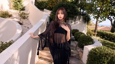 Woman in a sheer black K-pop–inspired outfit posed on a sunlit Mediterranean white-walled terrace with lush greenery — 5K Ultra HD desktop wallpaper.