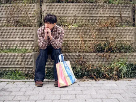 Cortis GREENGREEN 5K Ultra HD PC desktop wallpaper: a man in a plaid shirt sitting on concrete steps by a rainbow tote bag against an urban textured wall.