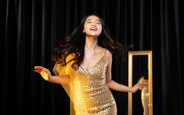 Cheerful young woman in a sparkling gold dress dancing before black curtains, light streaks and a reflected frame — 5K Ultra HD PC Desktop Wallpaper and Background.