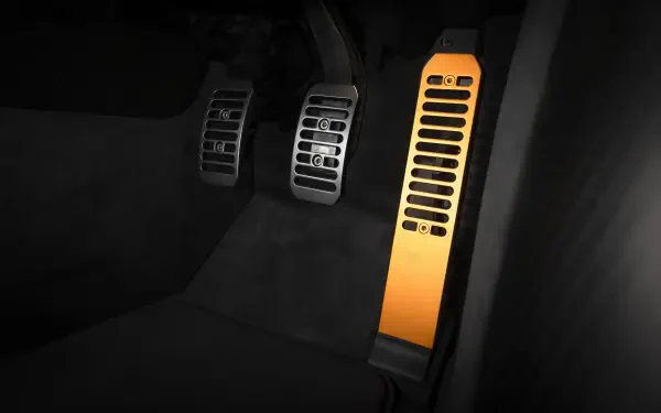 Close-up of McLaren McL 6GT driver footwell showing metal brake and clutch pedals and an illuminated orange dead pedal — 8K Ultra HD PC desktop wallpaper.