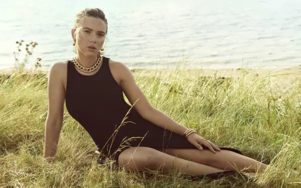 Person reclining in grassy shoreline wearing a black sleeveless dress and layered gold necklaces — 8K Ultra HD PC desktop wallpaper/background.