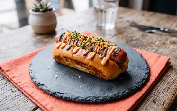 5K Ultra HD PC desktop wallpaper of a gourmet hot dog sandwich on a slate plate, drizzled with mustard, topped with relish and chives, set on a rustic wooden table.