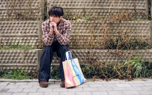 Cortis GREENGREEN 5K Ultra HD PC desktop wallpaper: a man in a plaid shirt sitting on concrete steps by a rainbow tote bag against an urban textured wall.