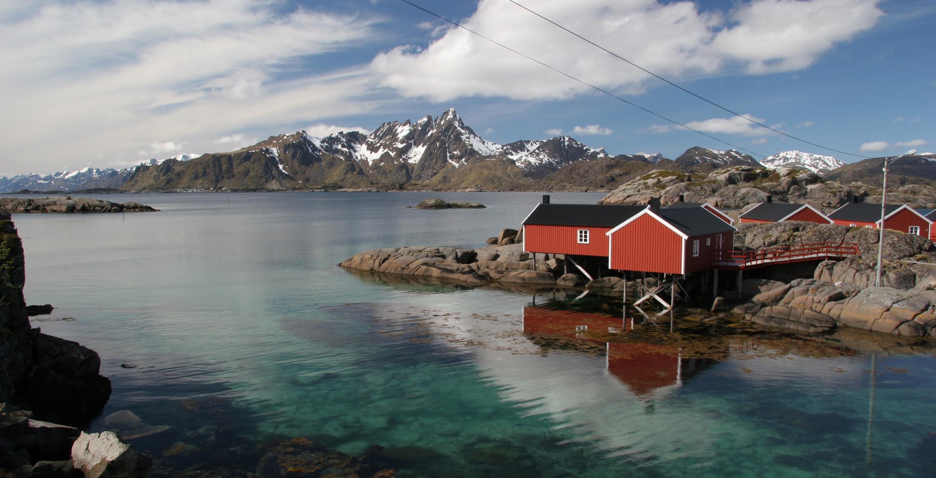 A serene view of a coastal town with red wooden houses beside clear blue waters, framed by majestic mountains and a bright sky, creating a captivating HD desktop wallpaper.