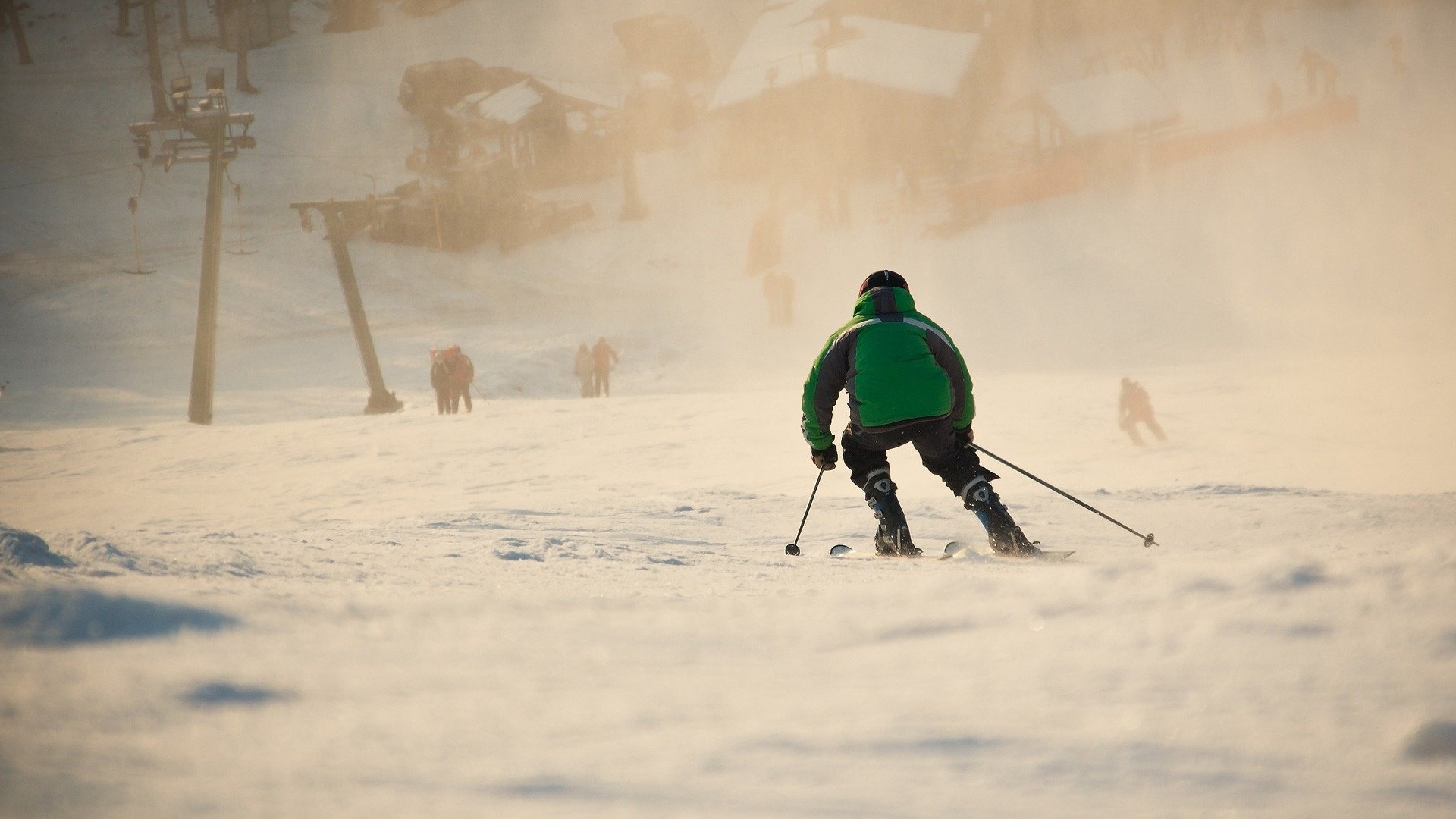 Skier in a green jacket carving through sunlit snow with a ski lift and distant figures — dynamic skiing sports scene, HD PC desktop wallpaper and background.