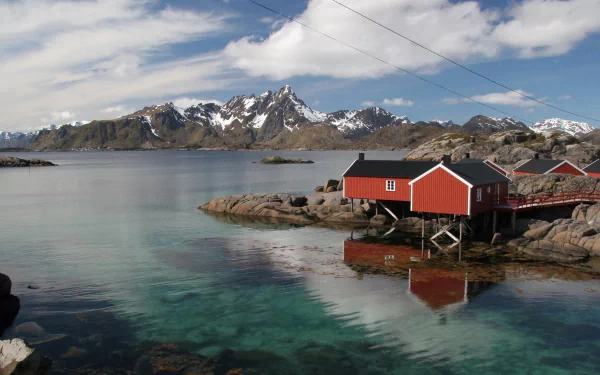 A serene view of a coastal town with red wooden houses beside clear blue waters, framed by majestic mountains and a bright sky, creating a captivating HD desktop wallpaper.
