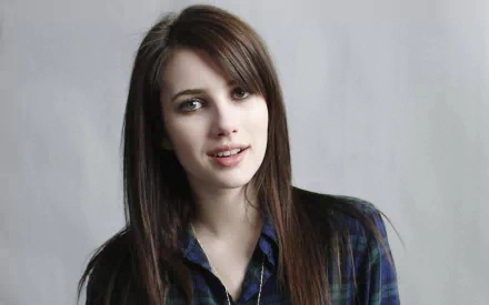 A close-up of celebrity Emma Roberts, featuring her with long hair and a plaid shirt, set against a soft gray background. An HD desktop wallpaper capturing her engaging expression.