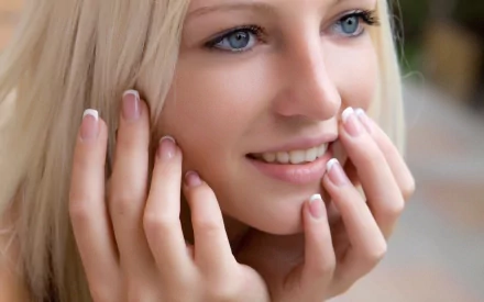 A pretty woman with blonde hair smiles gently, her hands framing her face, creating a serene and inviting atmosphere for an HD desktop wallpaper background.