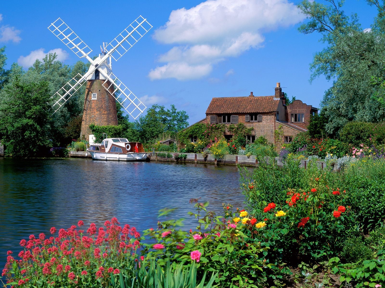 HD desktop wallpaper featuring a scenic man-made windmill beside a river, with colorful flowers and a charming house under a bright blue sky.