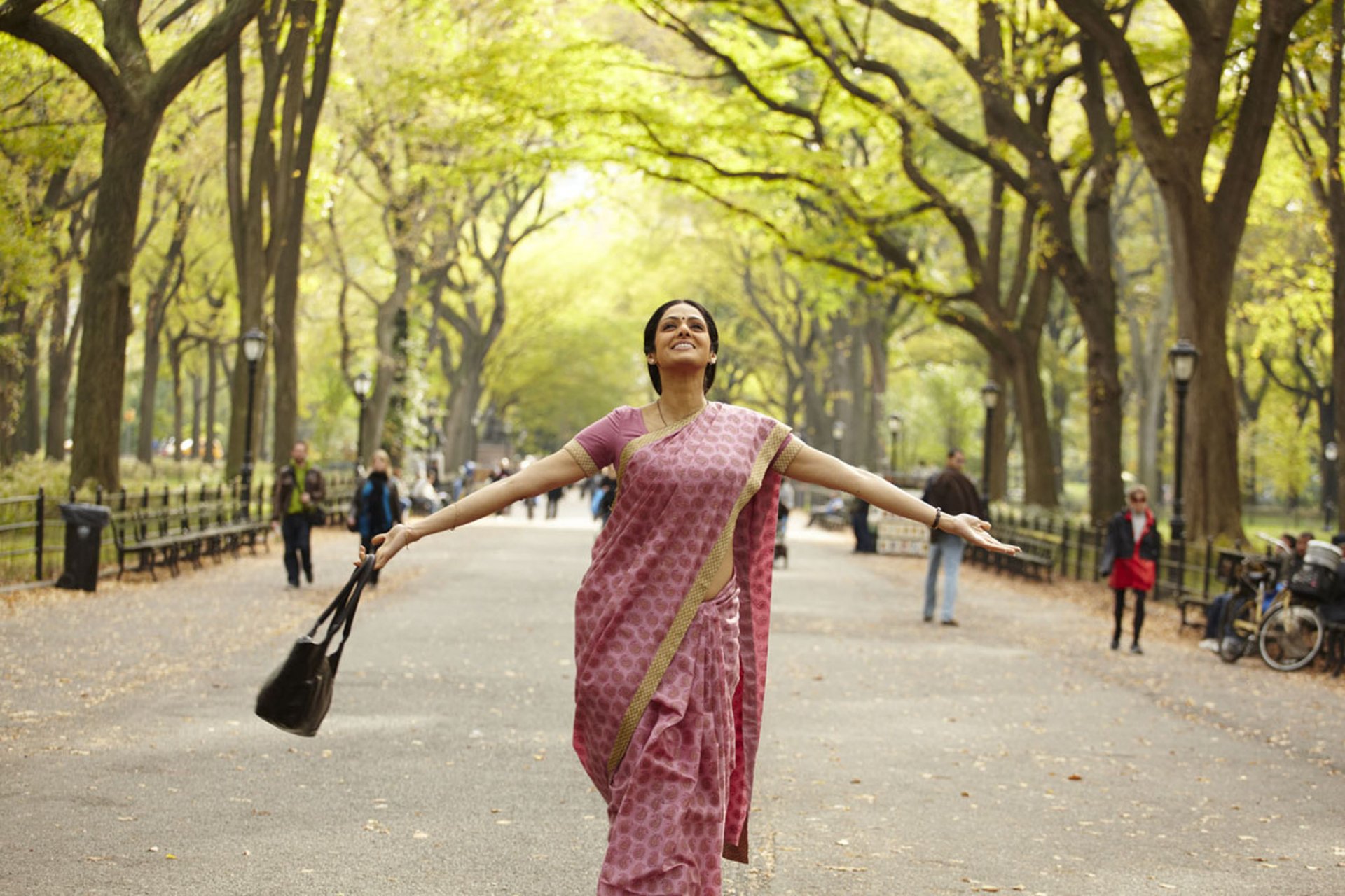 HD PC desktop wallpaper: joyful woman in a pink sari, arms outstretched, strolling down a sunlit, tree-lined park avenue.