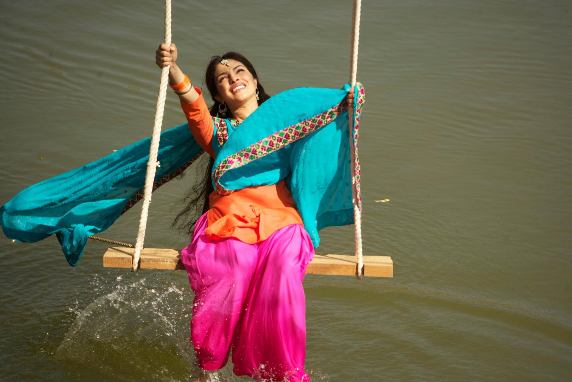 HD desktop wallpaper of celebrity Priyanka Chopra joyfully swinging over water, dressed in vibrant traditional attire with flowing turquoise scarf.