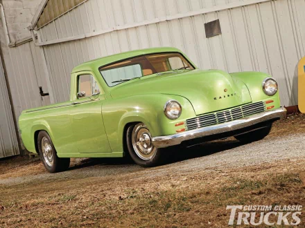 HD PC desktop wallpaper background: lime-green Kaiser pickup vehicle parked on gravel before a corrugated metal garage, classic chrome bumper and rounded fenders.