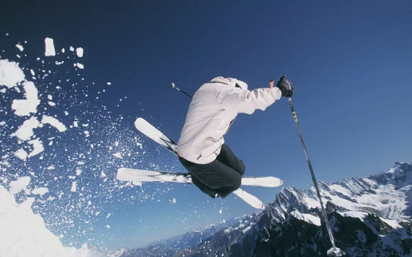 HD PC desktop wallpaper capturing a skier mid-air against a clear blue sky and snowy mountain backdrop, showcasing dynamic action in the sport of skiing.