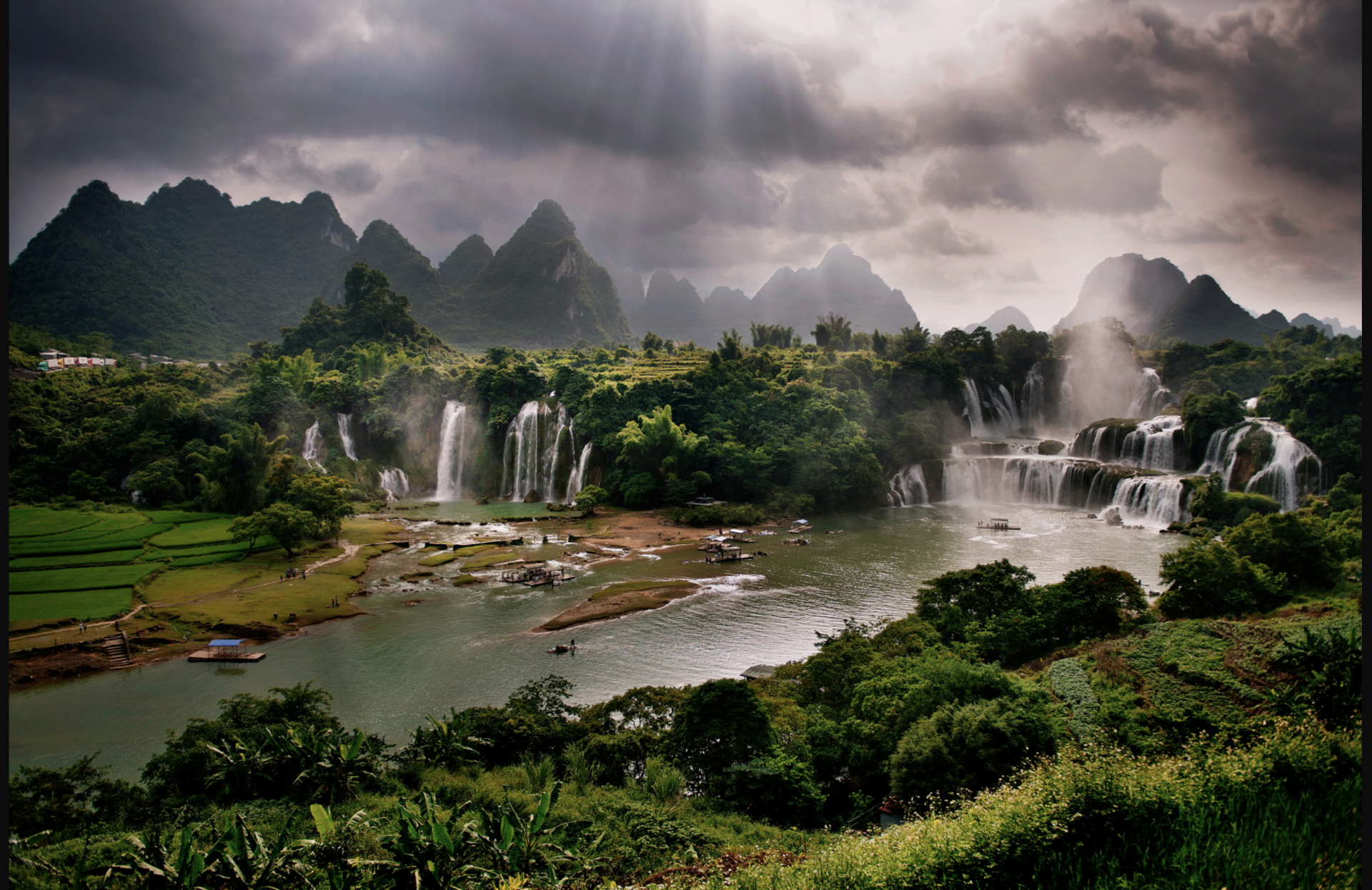 HD desktop wallpaper showcasing a breathtaking landscape of a majestic waterfall surrounded by lush greenery and dramatic mountains under a cloudy sky.