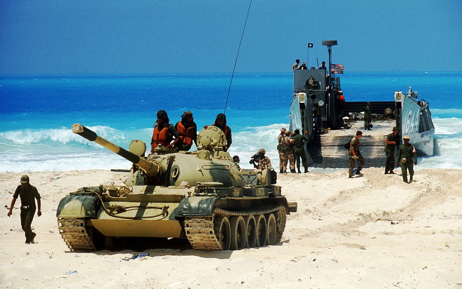 HD desktop wallpaper showing a military tank and soldiers on a sandy beach near a landing craft against a clear blue ocean backdrop.