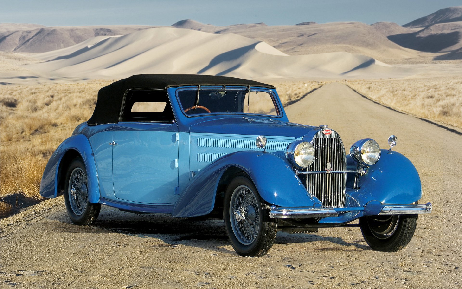 HD PC desktop wallpaper featuring a classic blue Bugatti vehicle parked on a desert road with sand dunes in the background.