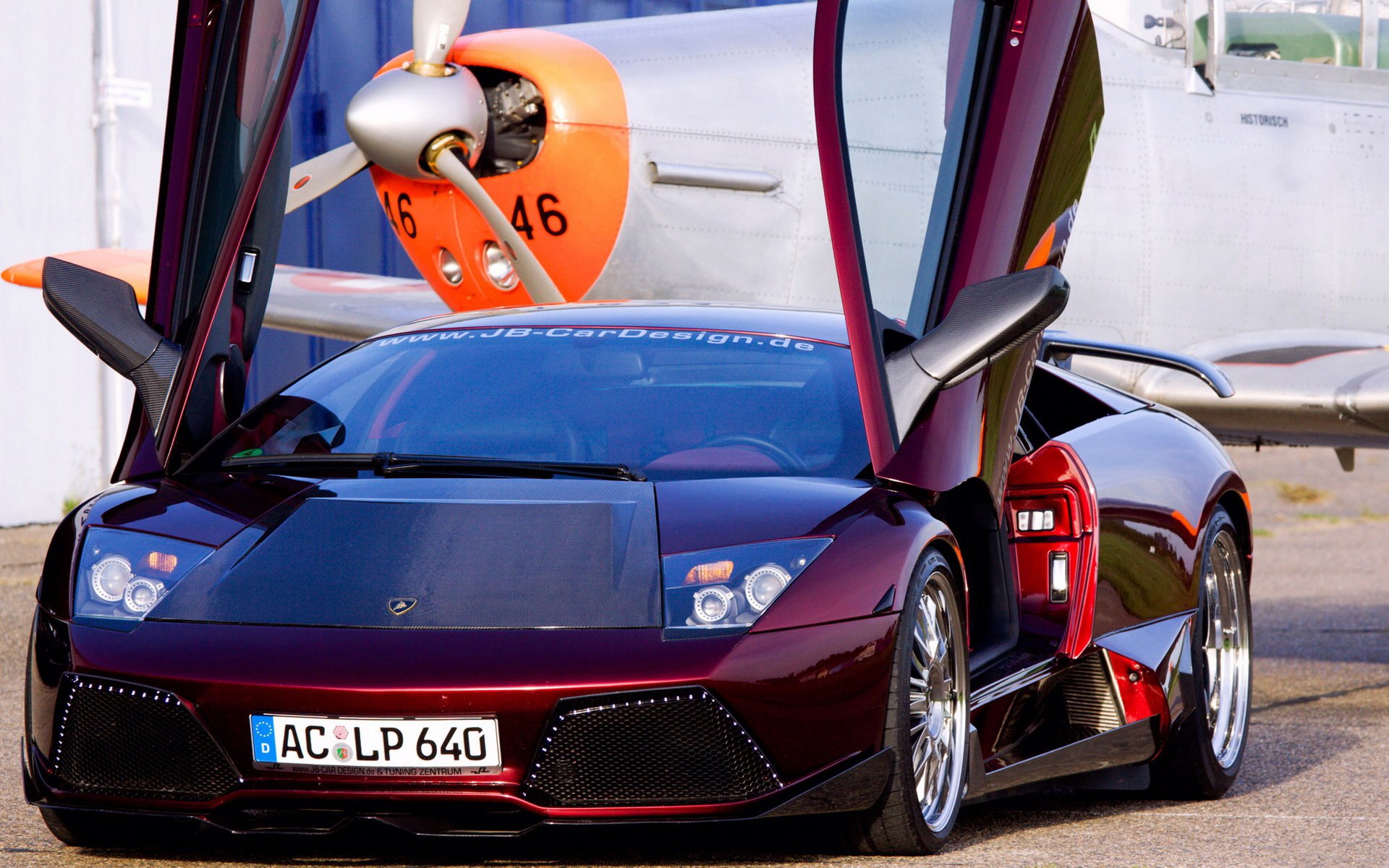 HD PC desktop wallpaper featuring a sleek Lamborghini Murcielago with its scissor doors open, parked in front of a vintage propeller airplane.