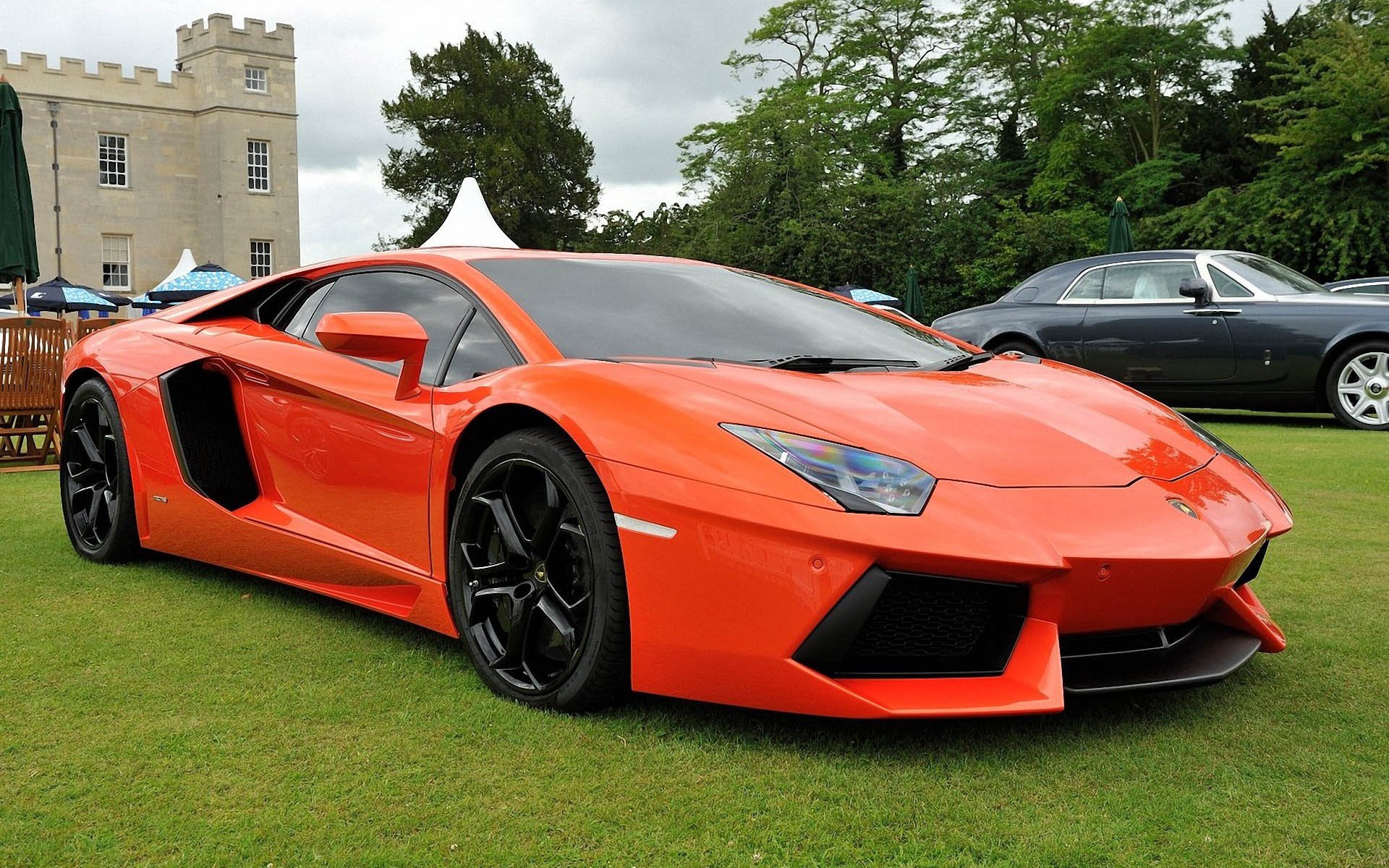 HD PC desktop wallpaper of a vehicle: orange Lamborghini Aventador LP 700-4 parked on a lawn in front of a manor, three-quarter front view with black wheels and sharp aerodynamic lines.