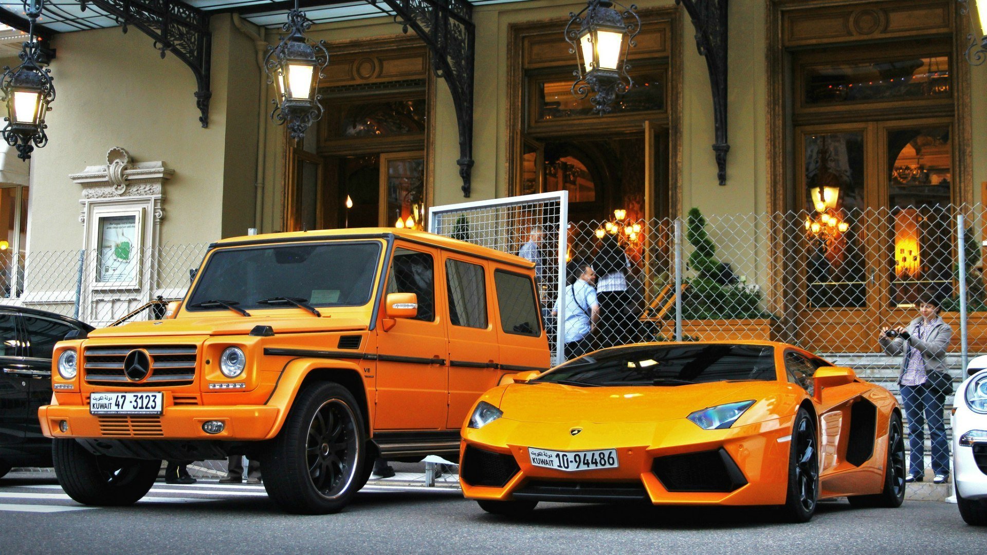 HD PC desktop wallpaper featuring a bright orange Lamborghini Aventador LP 700-4 parked beside an orange Mercedes G-Class SUV in front of a classic building.