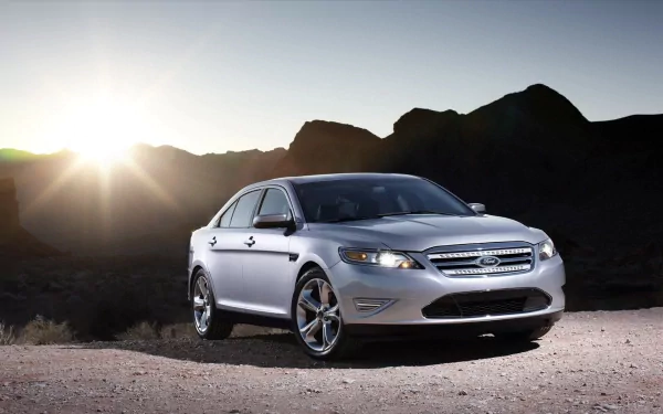HD PC desktop wallpaper: silver Ford Taurus vehicle parked on a desert road at sunset, mountains silhouetted behind and sun flare near the horizon.
