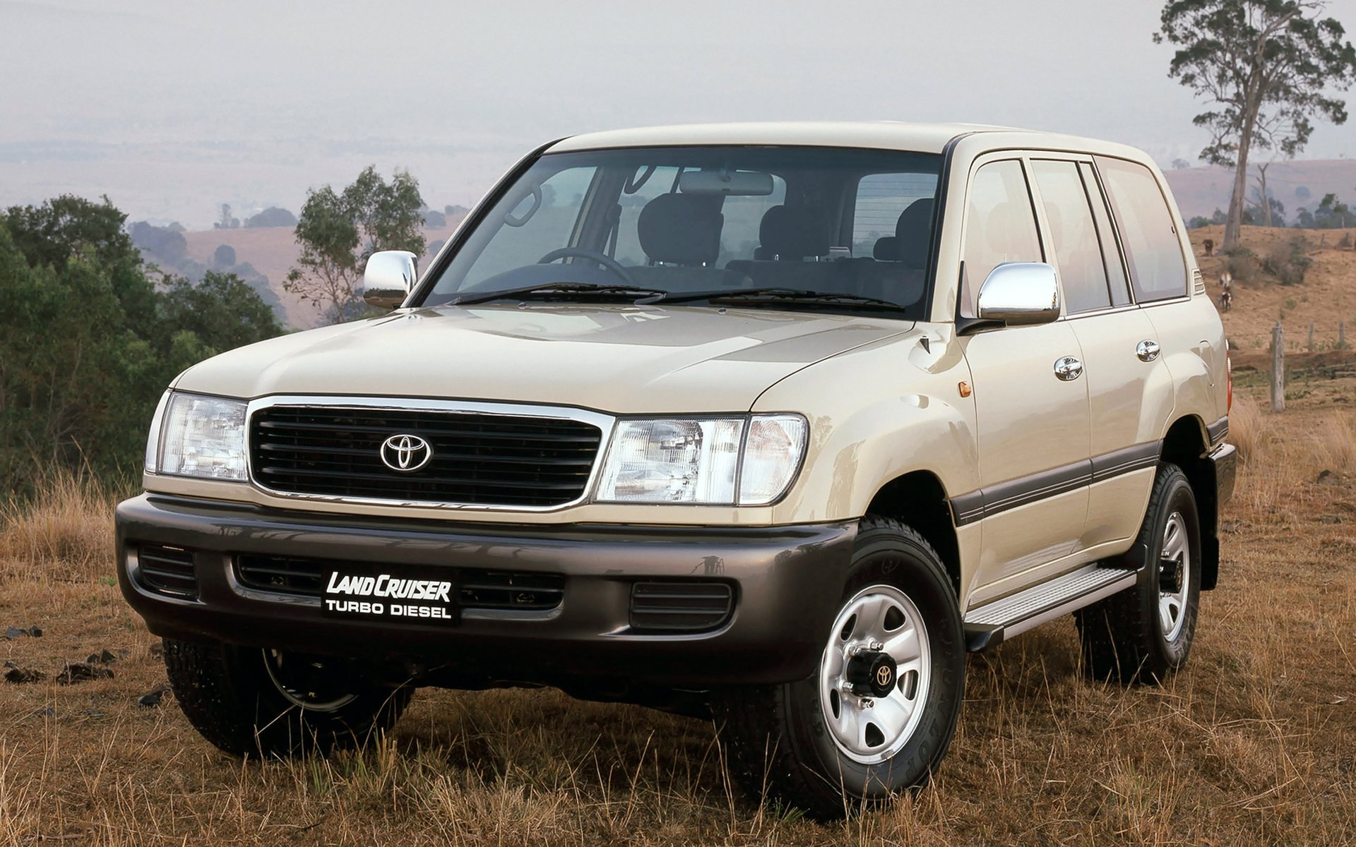 HD desktop wallpaper featuring a Toyota Land Cruiser SUV parked in a grassy field with trees and hills in the background.